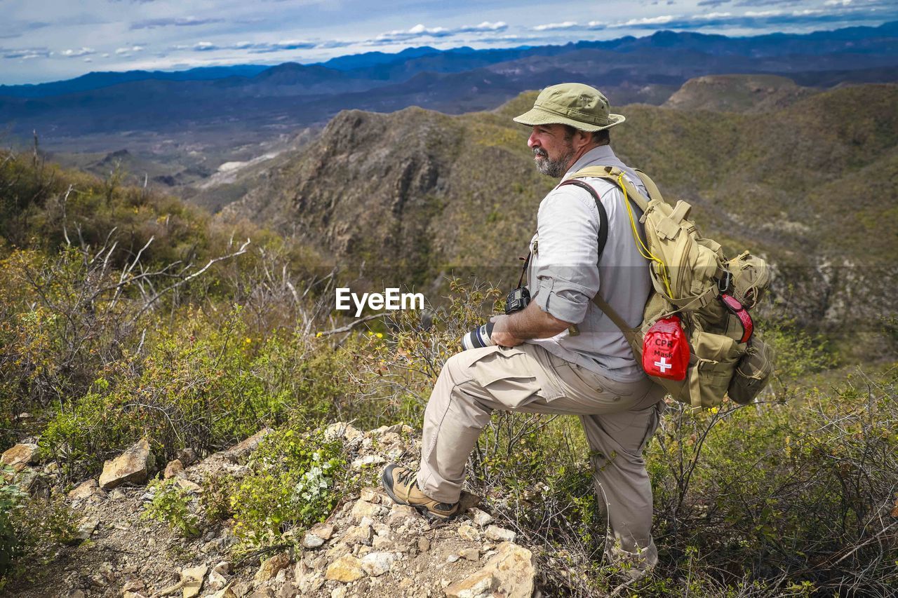 REAR VIEW OF MAN SITTING ON ROCK LOOKING AT MOUNTAIN