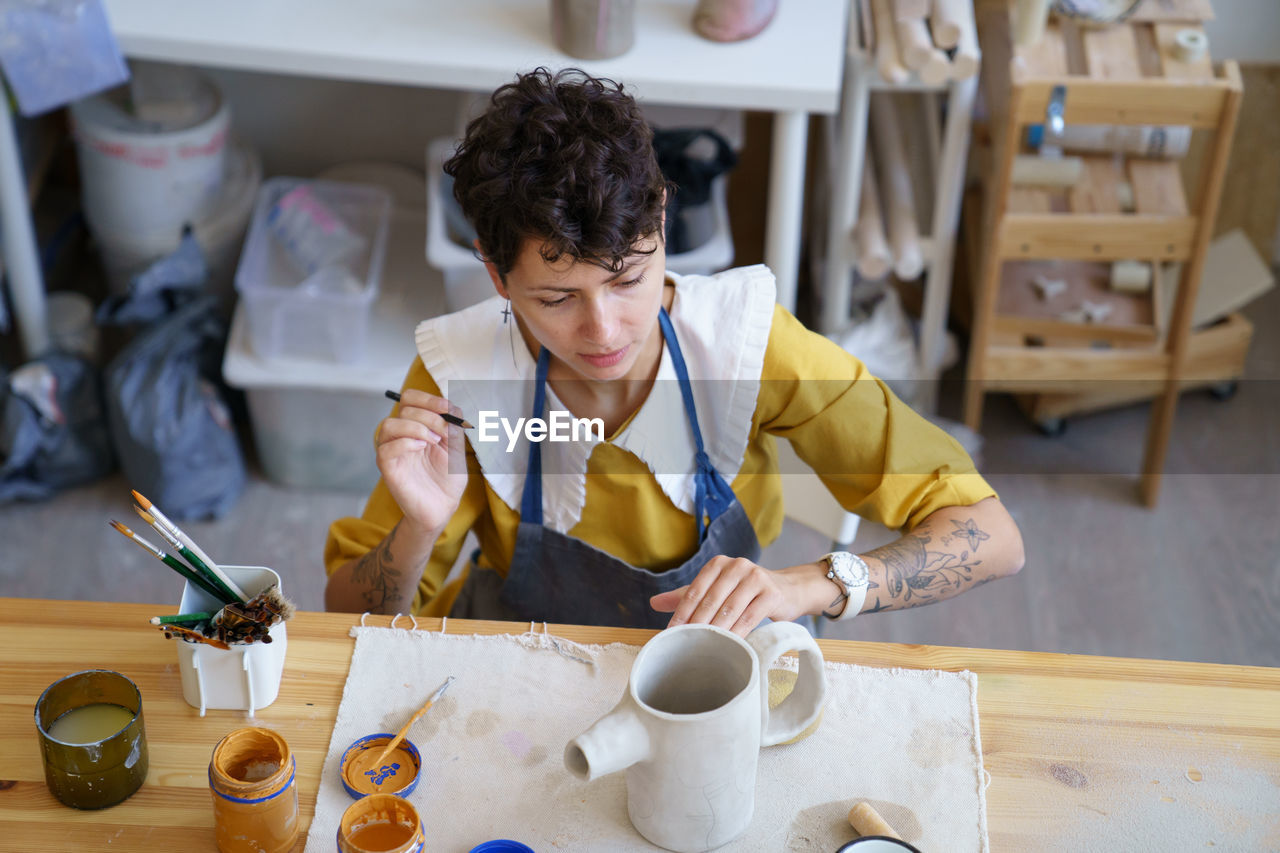 Woman artisian in pottery studio drawing on clay handmade teapot, female ceramist at work