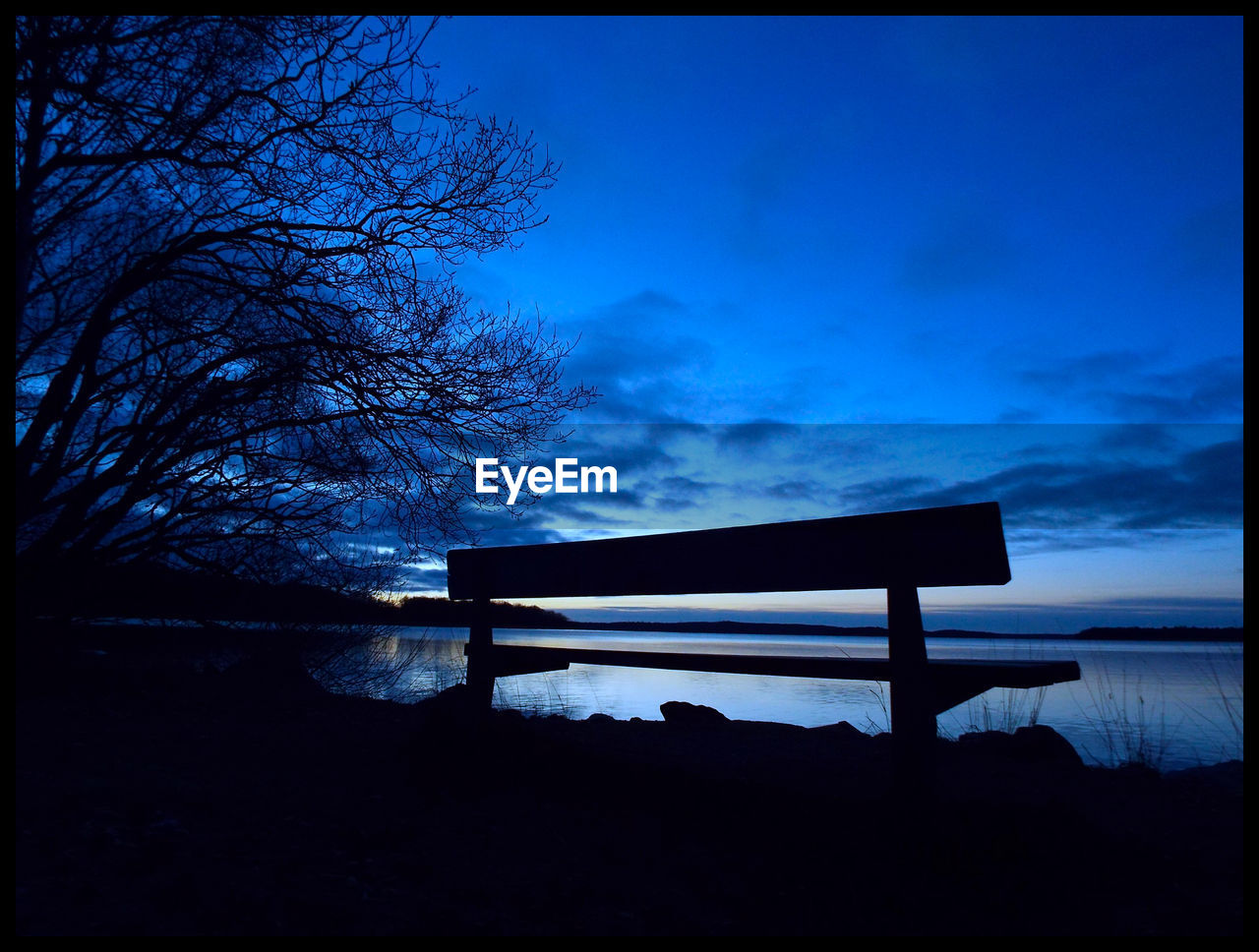 Silhouette bench by lake against blue sky