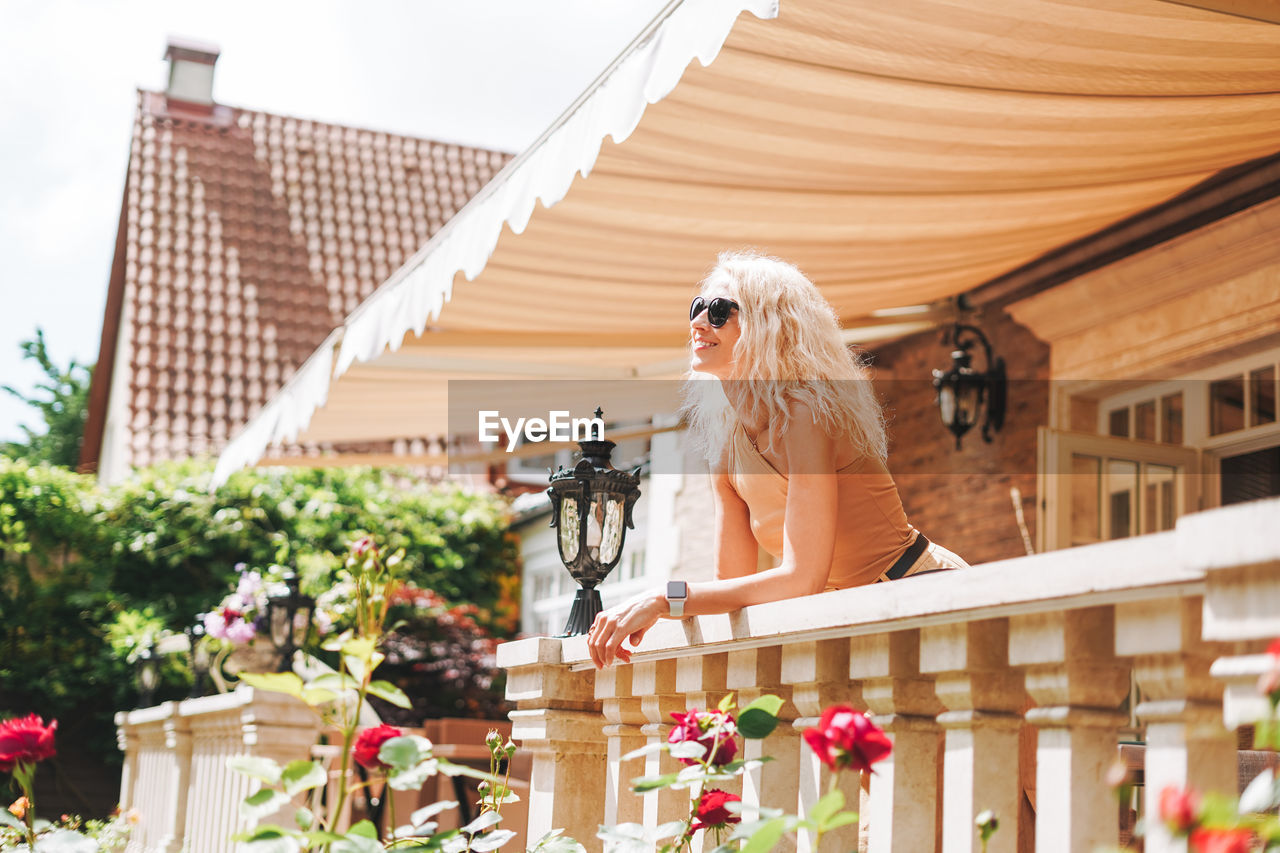 Adult elegant blonde woman in sunglasses beige clothes at house backuard, in summer city street cafe