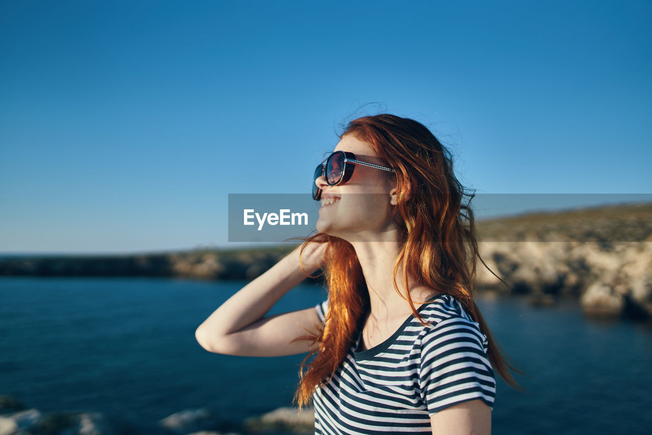 YOUNG WOMAN WEARING SUNGLASSES STANDING AGAINST SEA