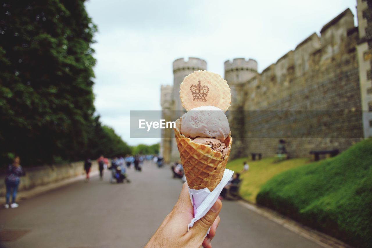 Hand holding ice cream cone against sky and castle fortified walls