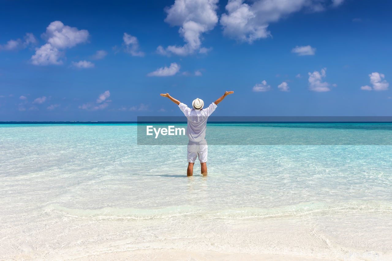 Rear view of man with arms raised standing in sea at beach against sky