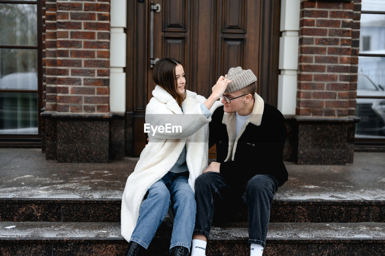  guy  and girl in are sitting on the porch of a brown brick house. girl puts  knitted hat 