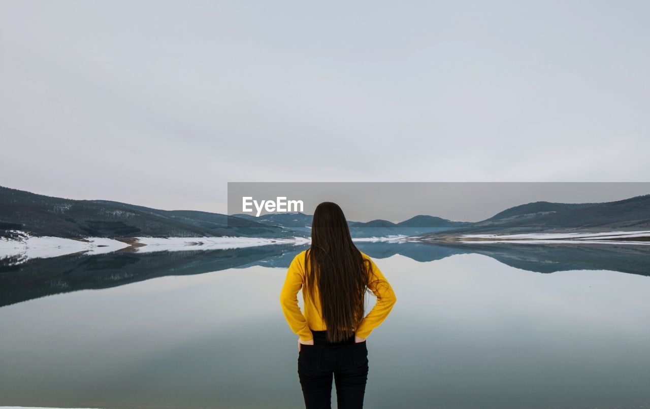 Rear view of woman with hands in back pockets standing by lake against clear sky