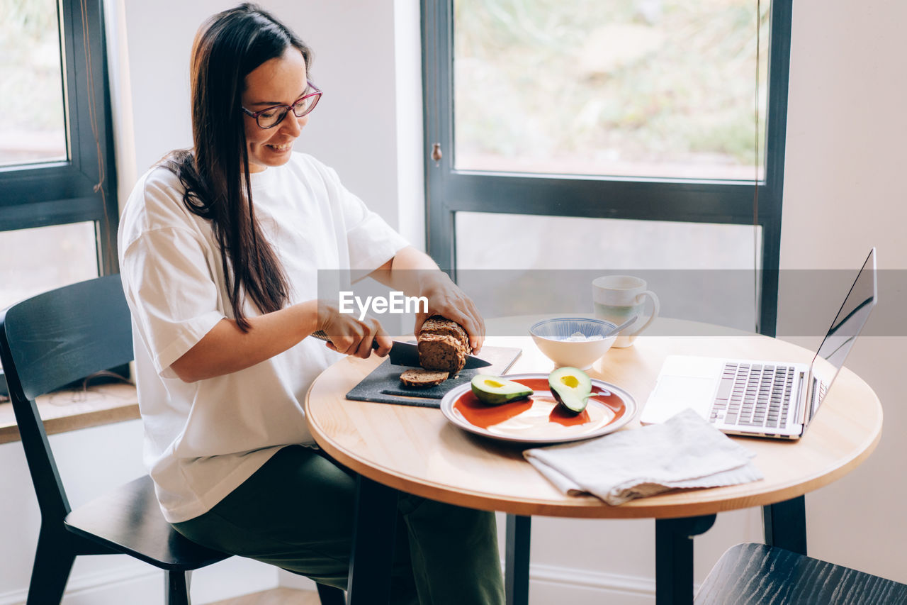 A young woman sitting at a table in front of a laptop makes a healthy breakfast.