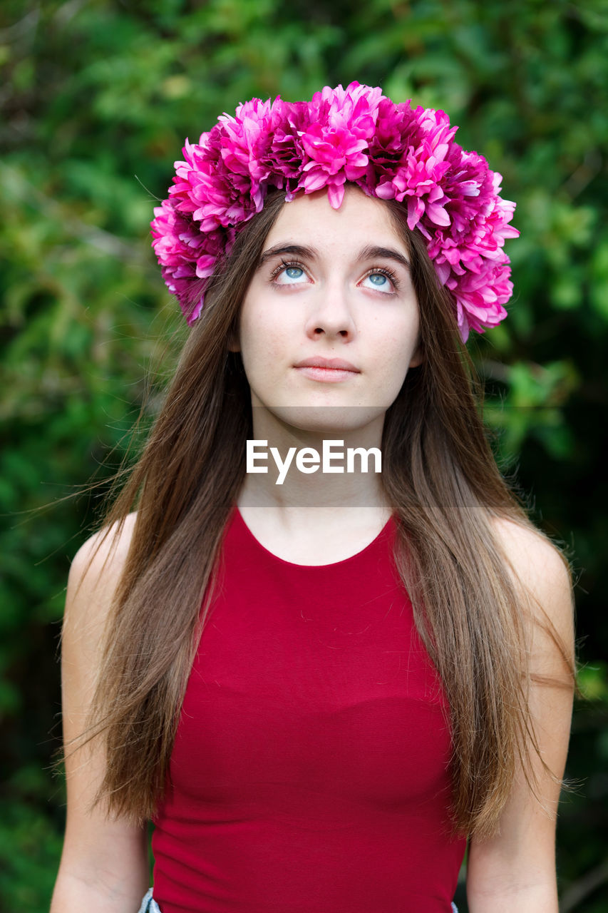 PORTRAIT OF BEAUTIFUL YOUNG WOMAN WITH PINK FLOWER STANDING AGAINST PLANTS