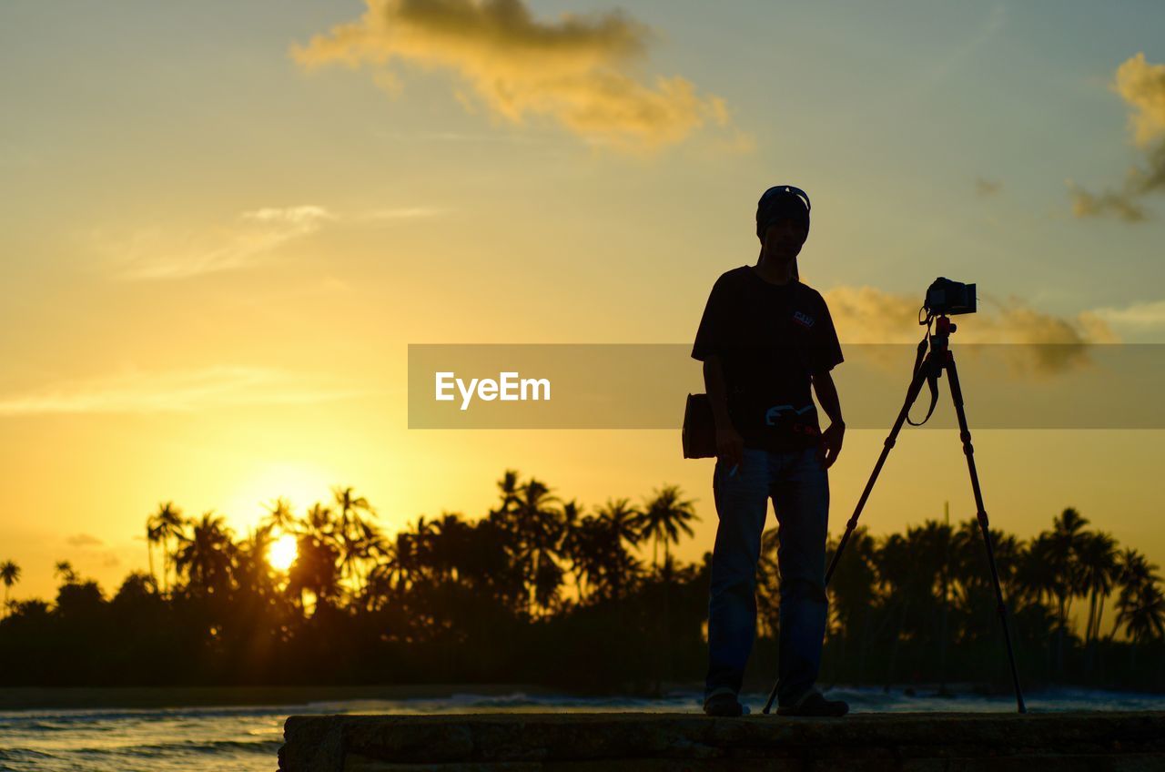 REAR VIEW OF SILHOUETTE MAN STANDING BY WATER AGAINST SKY