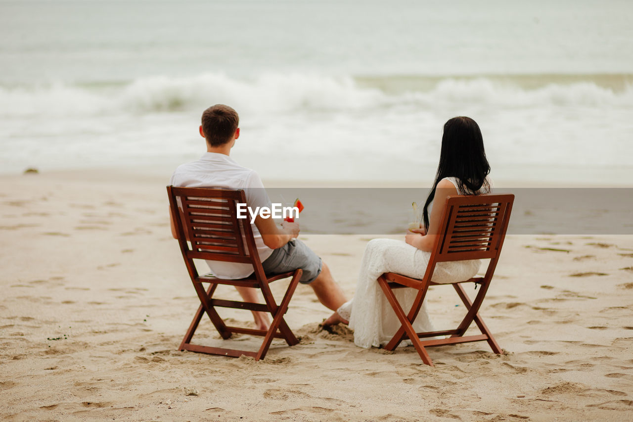 Rear view of couple having drink while sitting on chairs at beach