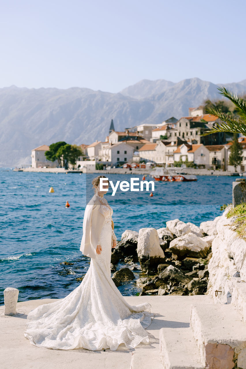 WOMAN STANDING BY SEA AGAINST MOUNTAINS