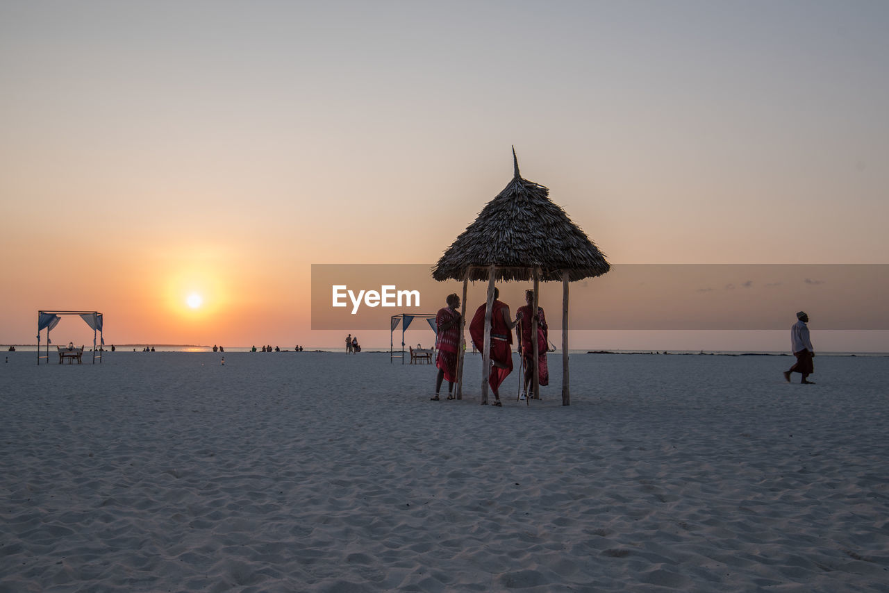 People standing in thatched roof at beach during sunset