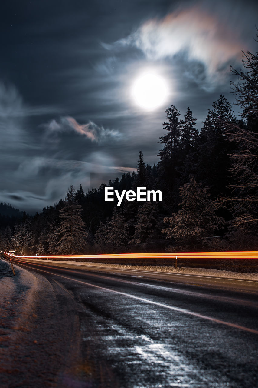 Light trails on road by trees against sky at night