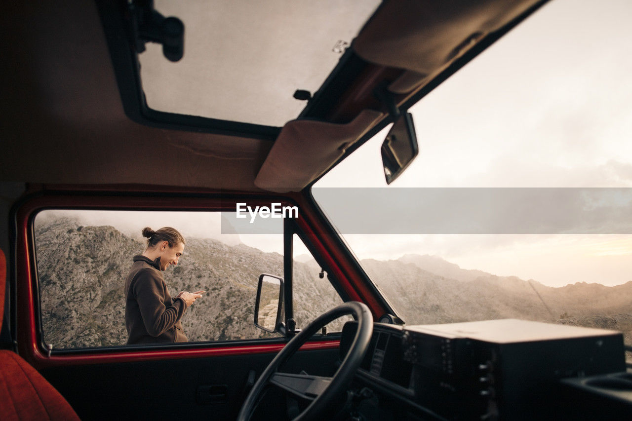 Side view of smiling man using smart phone seen from van window on vacation