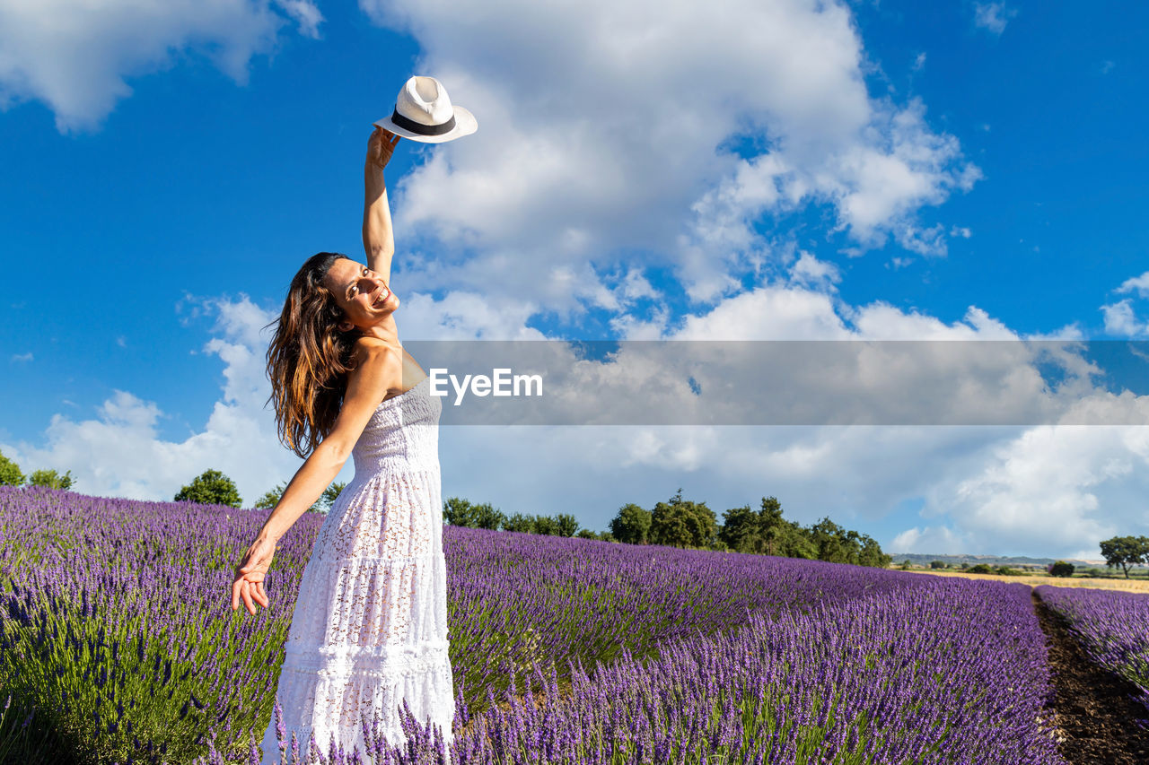 Young happy woman dances in a lavender field and raises her hat to the blue sky. 