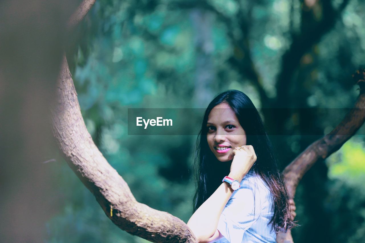 PORTRAIT OF SMILING YOUNG WOMAN STANDING AGAINST TREES