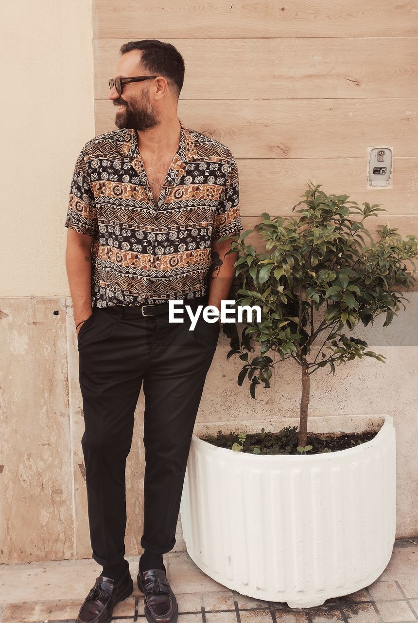 Young man looking away while standing on potted plant against wall