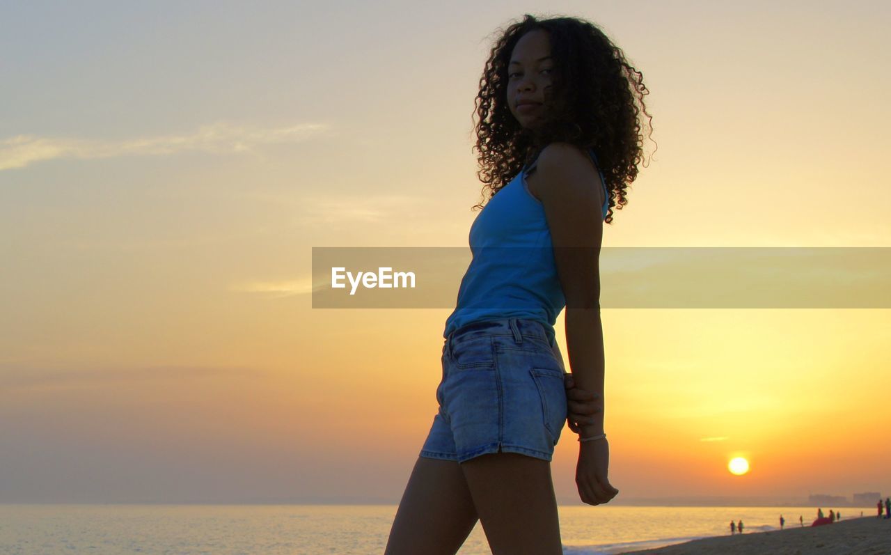 Woman standing by beach against sky during sunset