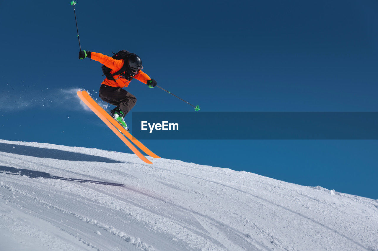 A skier in an orange jacket and a black helmet makes a jump on a sunny day against a clear sky. copy