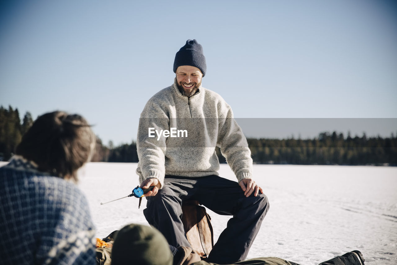 Happy mature man holding fishing rod sitting with friends during sunny day in winter