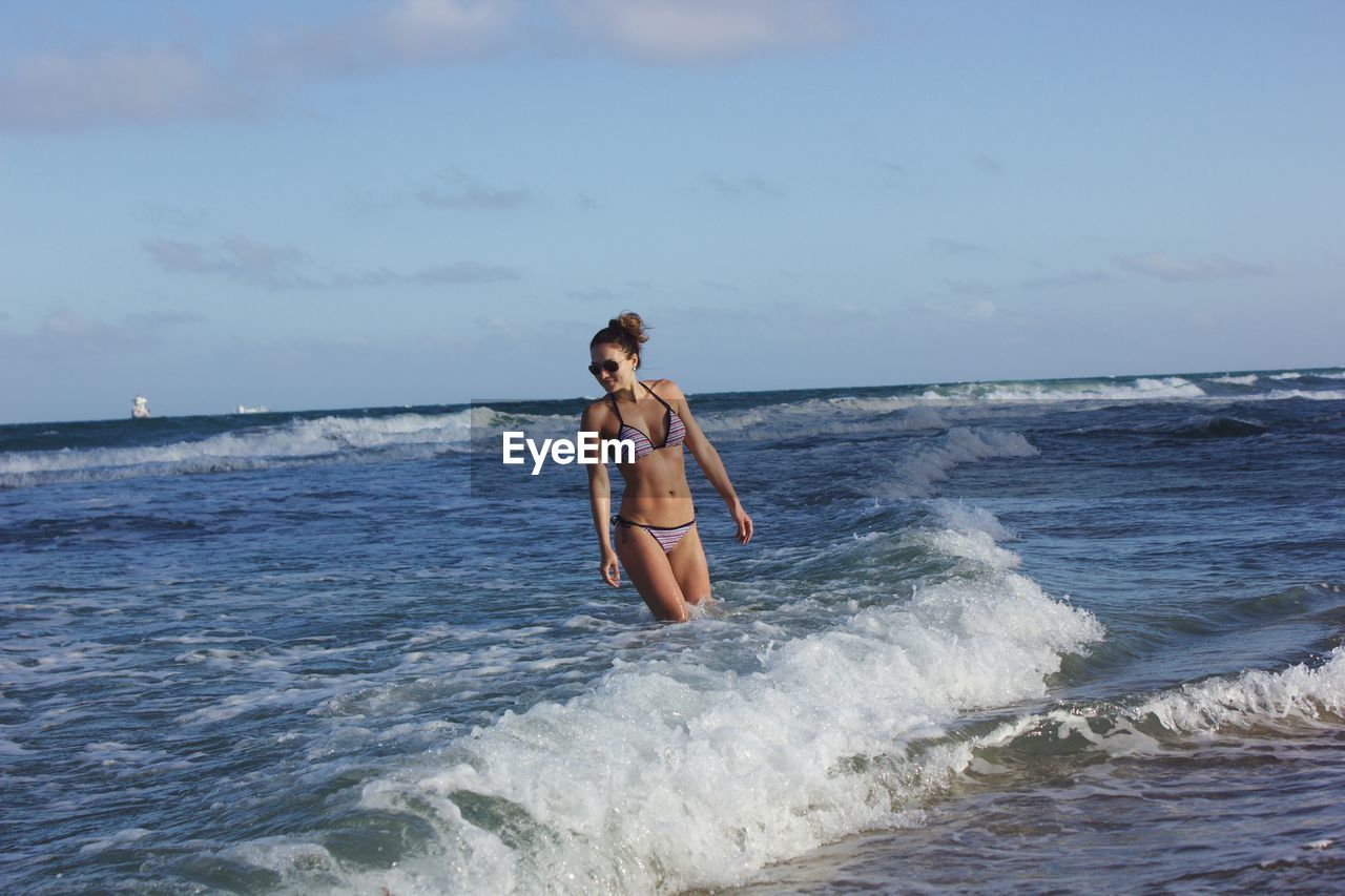 Young woman wearing bikini while standing in sea against sky