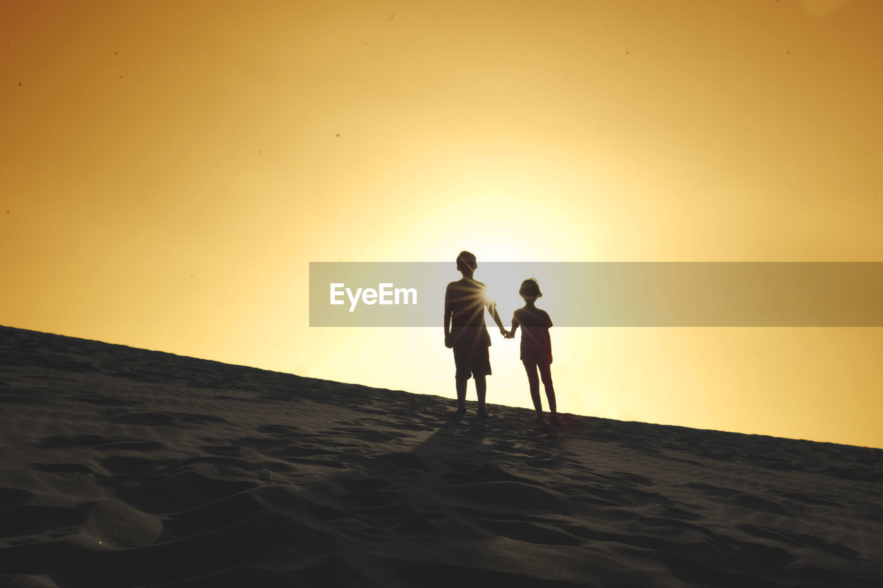 Silhouette siblings standing on sand against sky during sunset