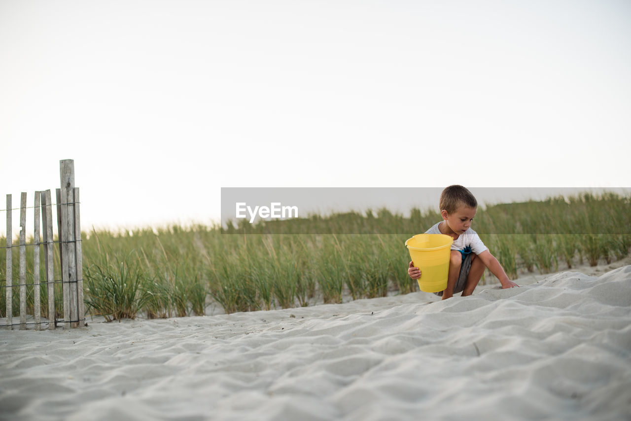 Young boy digging in sand on the beach with a yellow bucket at sunset