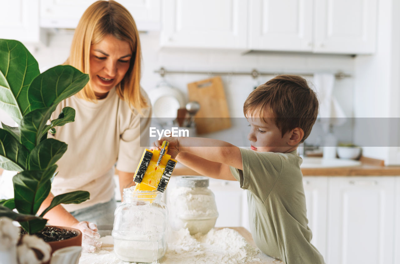 Young woman mother and her toddler boy son have fun while cooking with flour at table in kitchen 