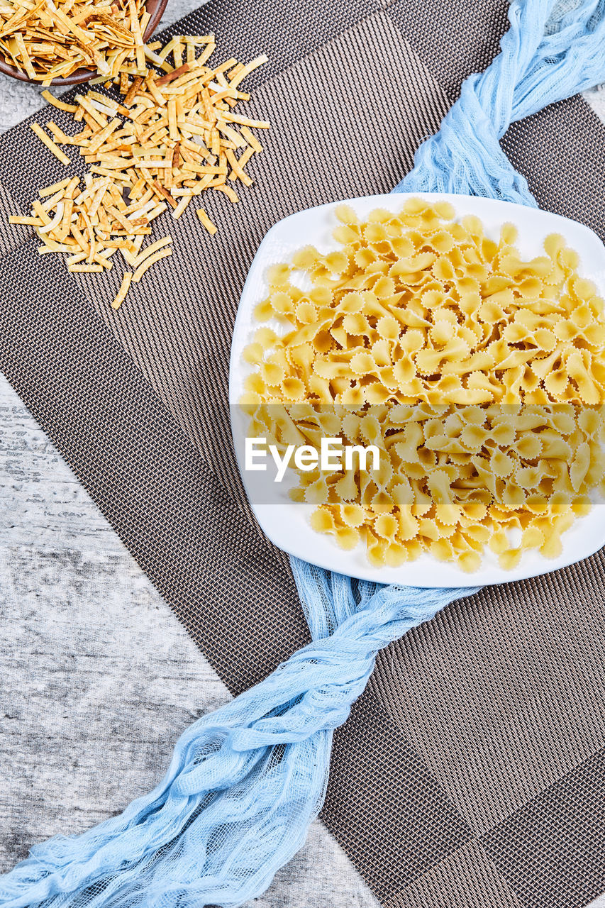 Two bowl of uncooked pasta with blue tablecloth on grey background