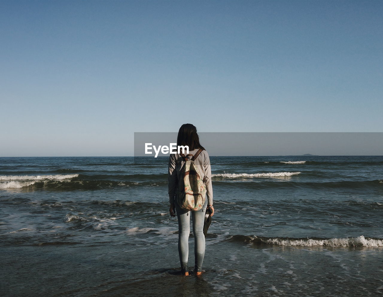 Full length rear view of woman with backpack standing on beach against clear sky