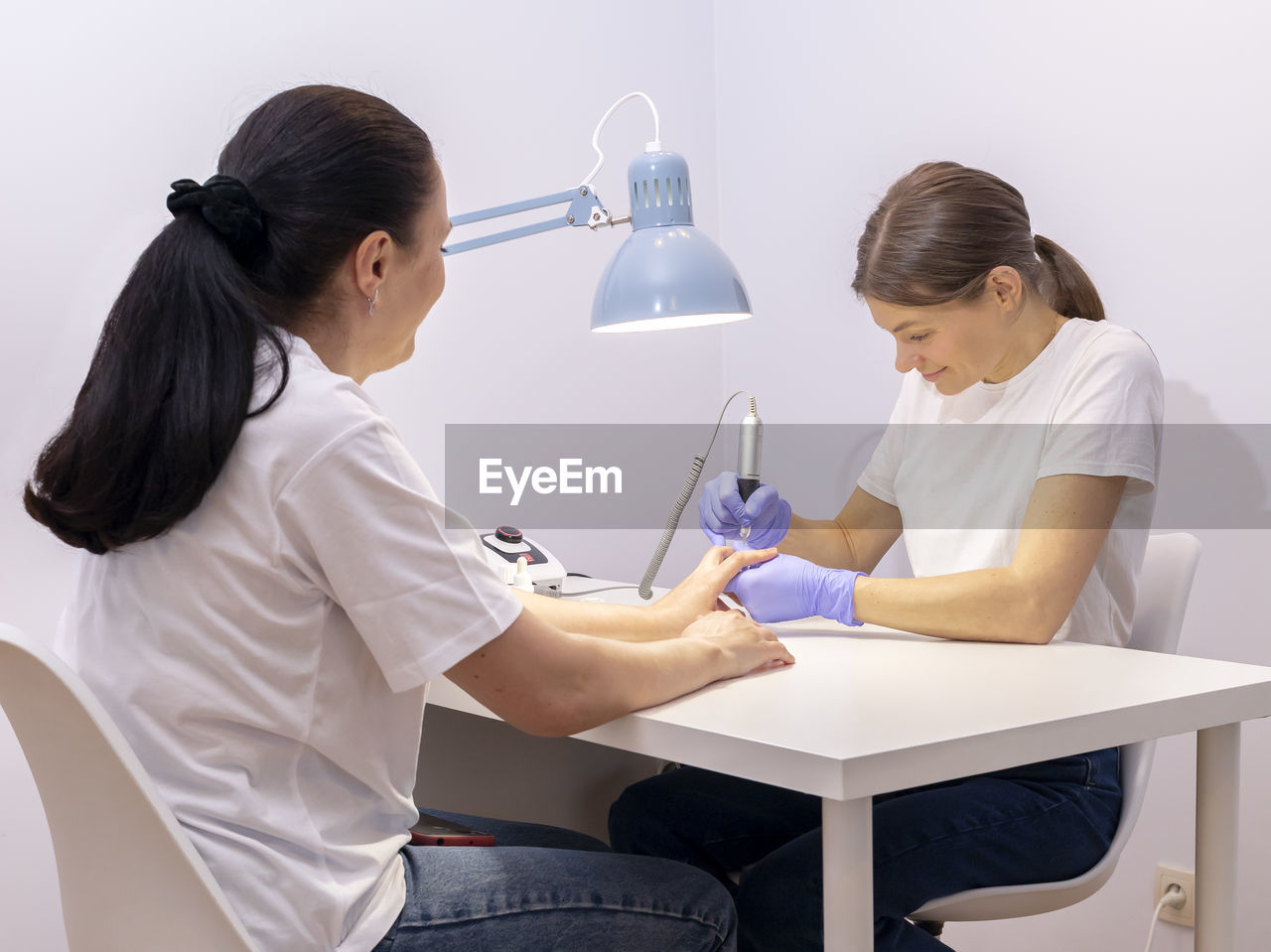 White nail technician in mask drills, removes mature client cuticle with hardware machine. customer