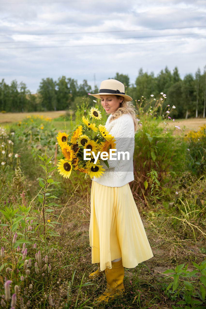 A cute girl in a brimmed hat and yellow rubber boots holds a bouquet of sunflowers in a field