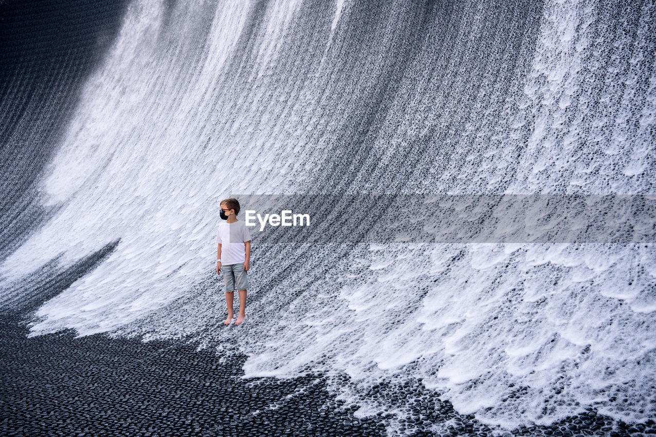 A boy in denim shorts and a white t-shirt looks at an artificial waterfall. 