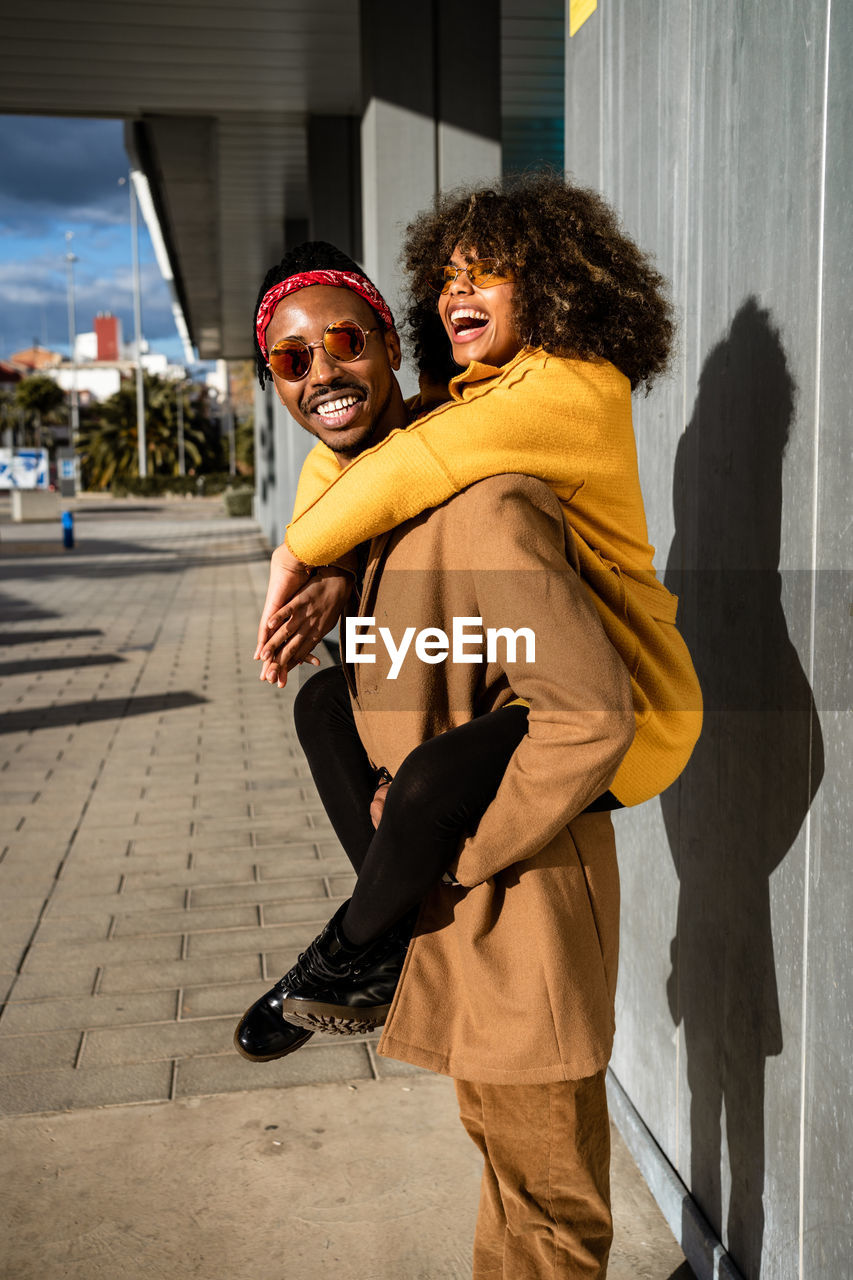 Cool african american man in sunglasses and coat giving piggyback ride to bright laughing girlfriend with afro curly hair spending time on street