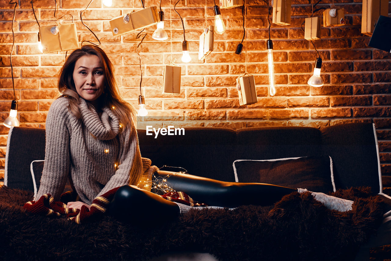 PORTRAIT OF YOUNG WOMAN SITTING AGAINST ILLUMINATED BRICK WALL IN ROOM