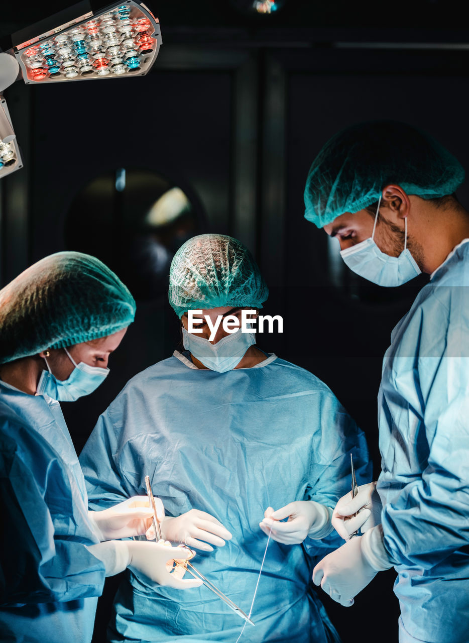 Group of focused professional surgeons with surgical tools and thread finishing operation of patient in operating theater