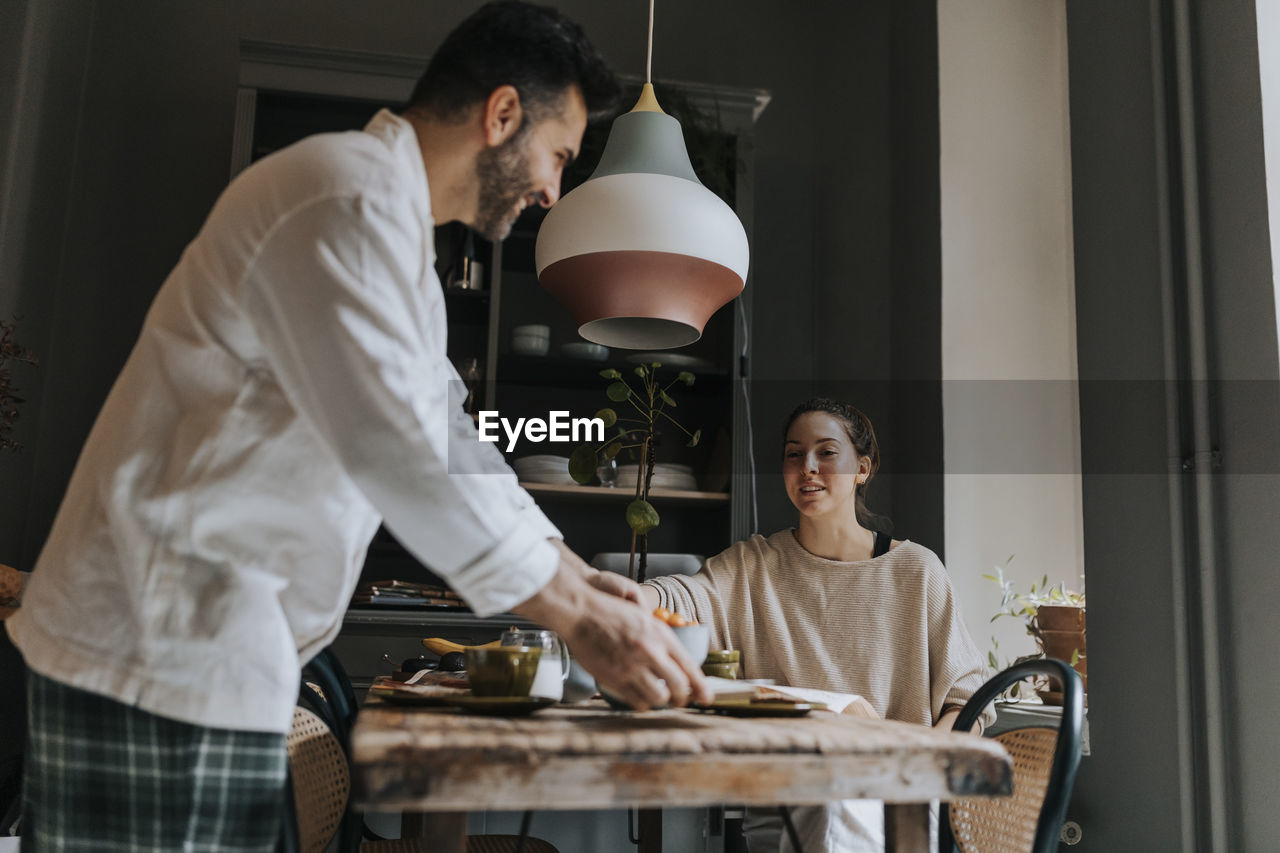 Smiling man serving breakfast with non-binary person sitting at dining table in home