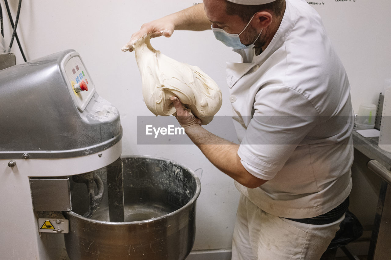 Mid adult chef preparing dough for bread by electric mixer in bakery during covid-19