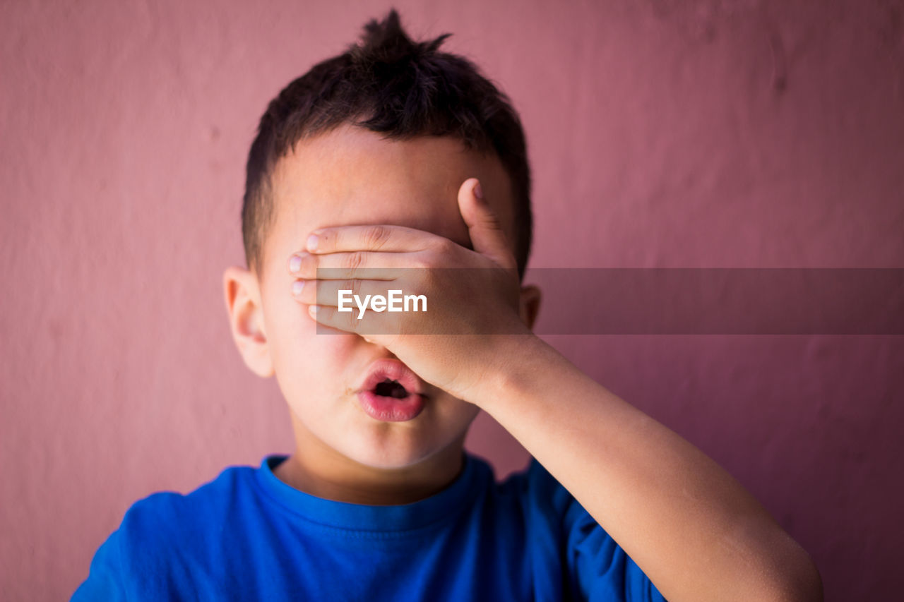 Close-up of boy with hands covering eyes against wall