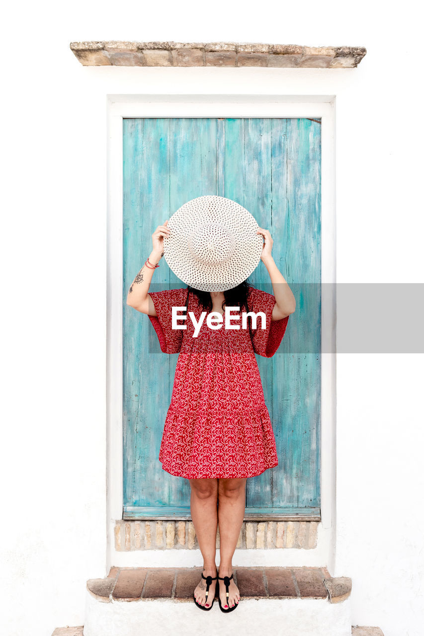 Anonymous female traveler in trendy apparel covering face with straw hat near aged building in greece