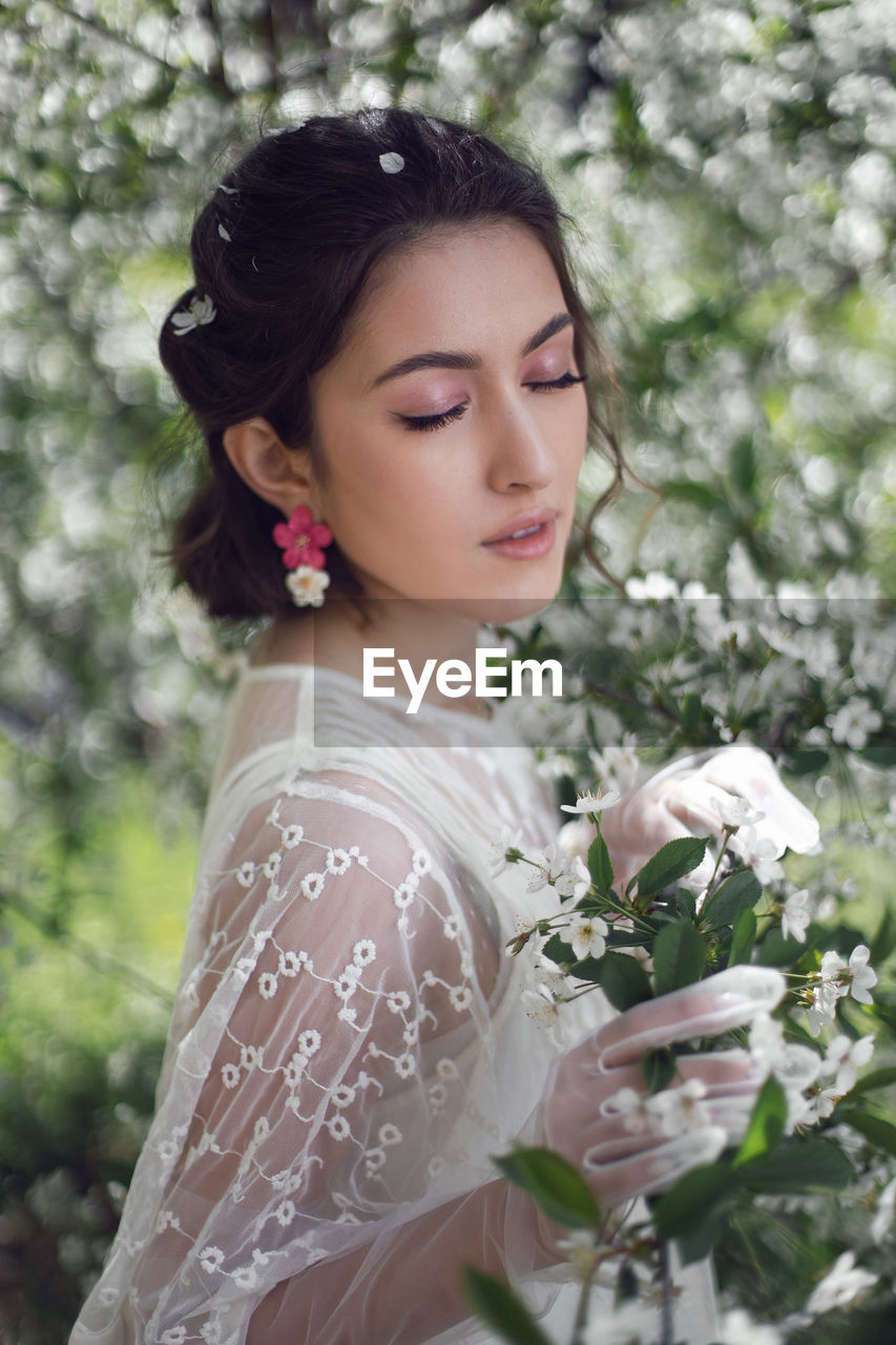 Portrait of a young beautiful woman in white clothes standing next to blooming cherry tree in spring