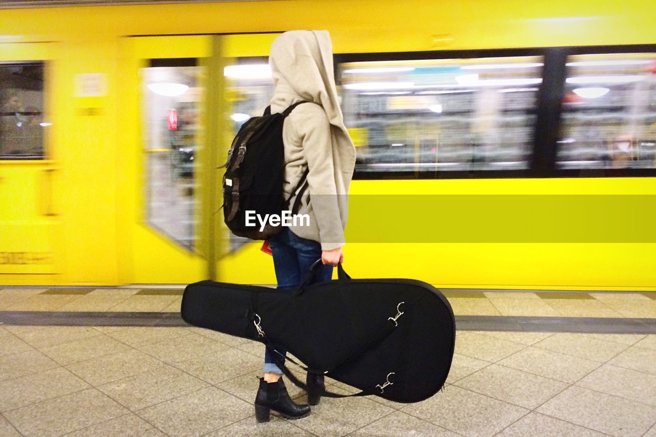 Side view of woman in hooded jacket holding guitar while waiting at railway station