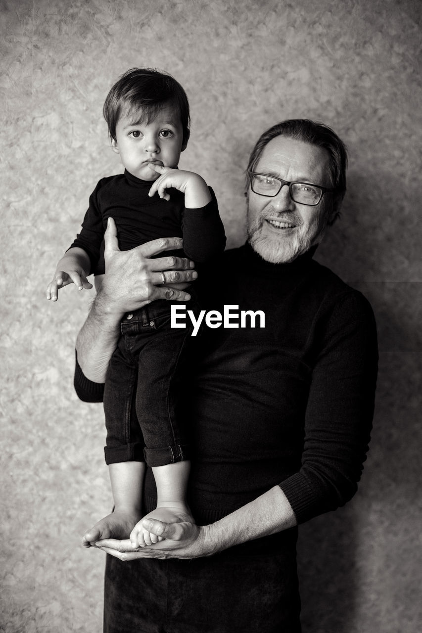 Grandfather with beard and glasses with his grandson sitting on his hands in the studio