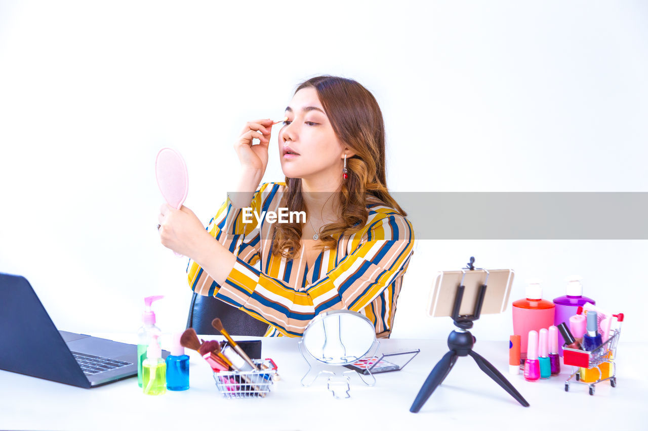 Young woman applying beauty product while blogging on table against white background