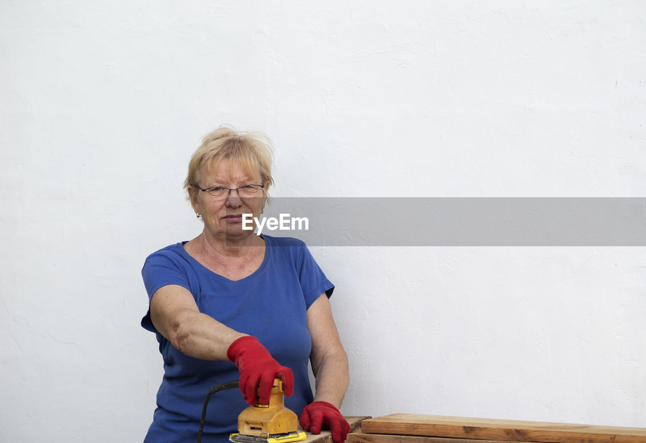 Senior caucasian woman in a blue t-shirt and red gloves is sanding a board with a sander 