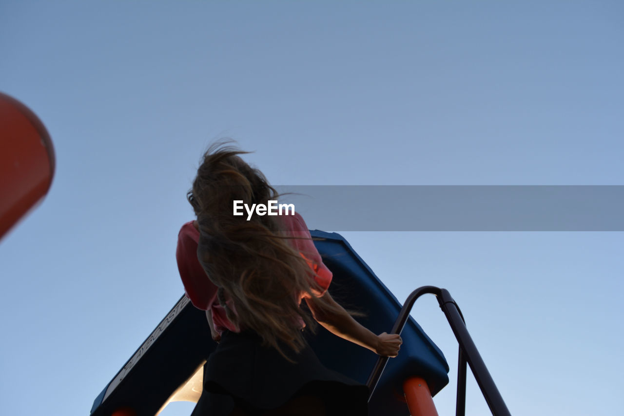 Low angle view of teenage girl with long hair standing on outdoor play equipment against clear sky