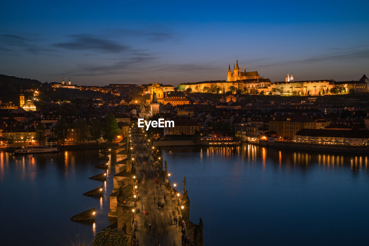 Illuminated buildings in city by river at dusk