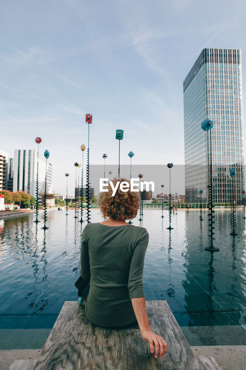 Rear view of woman sitting by lake in city against sky