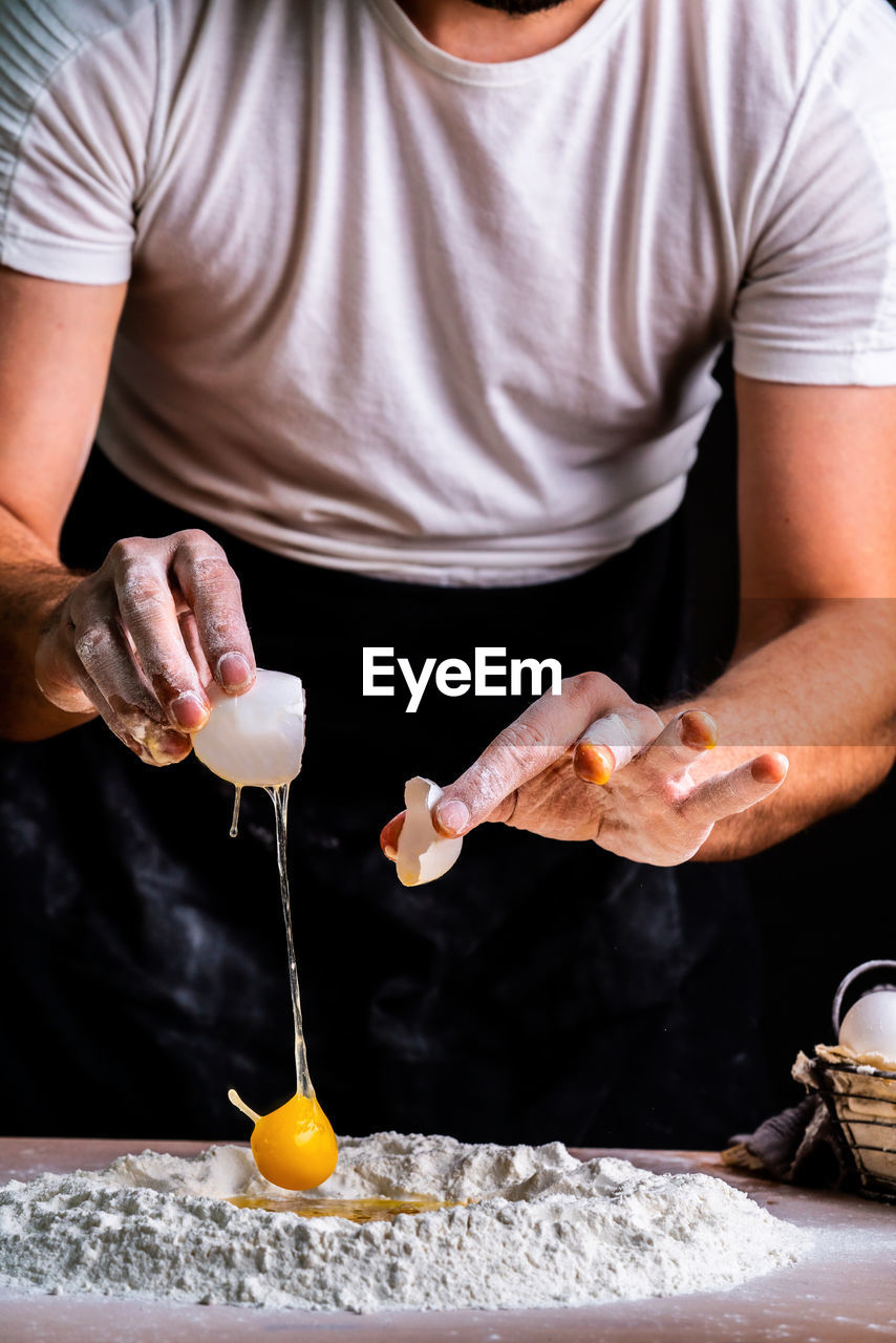 Faceless female baker breaking raw chicken egg into sifted flour pile on table cooking bread  person