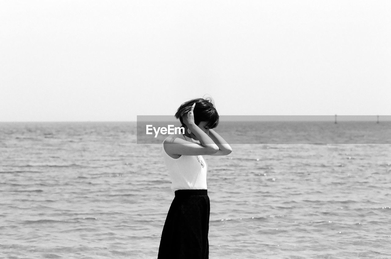 Side view of young woman standing at beach against sky during sunny day