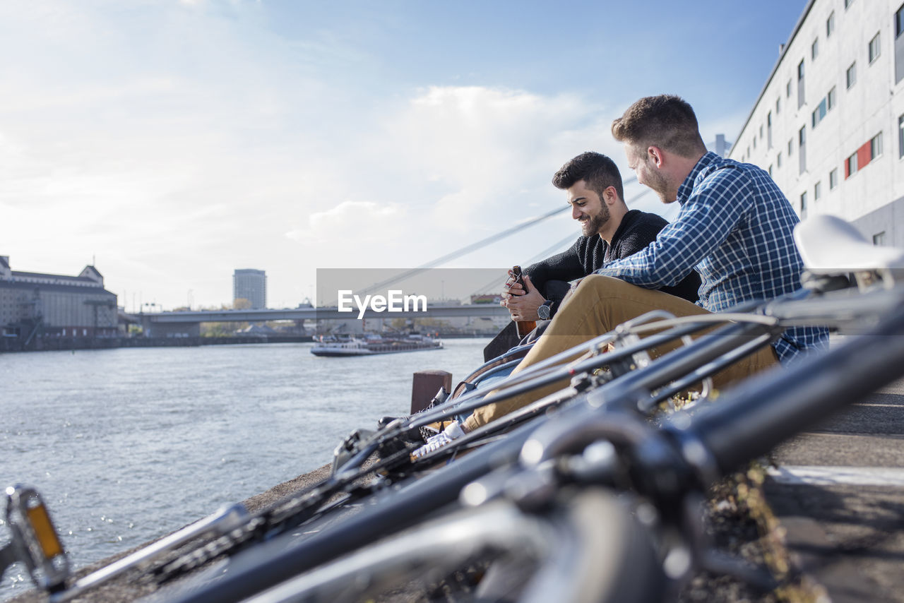 Friends sitting by bicycle in front of canal against clear sky